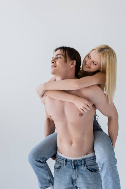 Smiling blonde woman hugging shirtless boyfriend in jeans isolated on grey  - 写真・画像