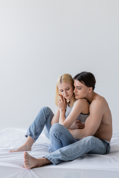 gespierde man knuffelen blond vriendin in jeans op bed geïsoleerd op grijs  - Foto, afbeelding
