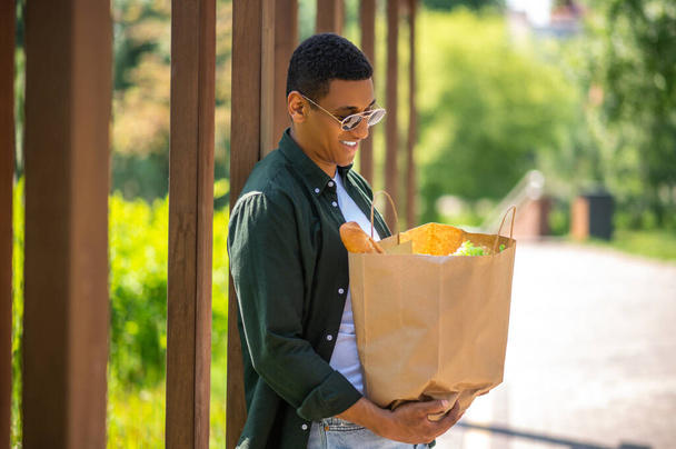 Anticipazione. Sorridente ragazzo dalla pelle scura in occhiali da sole guardando in borsa di cibo in piedi nel parco nella giornata di sole - Foto, immagini