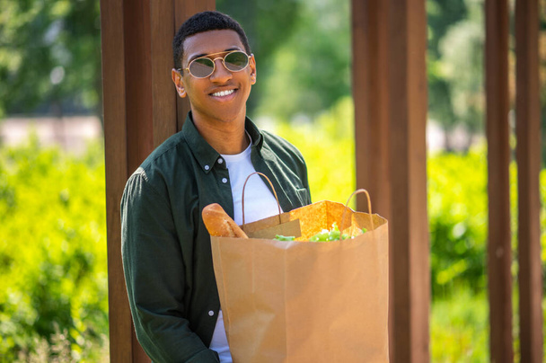 Excellent day. Young dark-skinned man in sunglasses smiling at camera holding food bag standing in park on sunny day - Photo, image