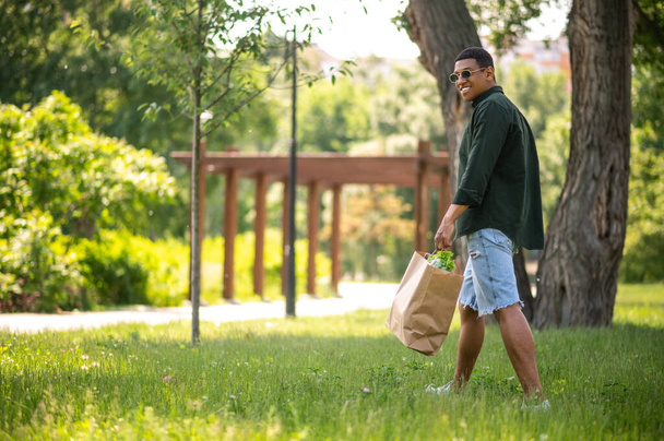 Walk in nature. Smiling dark-skinned guy in sunglasses walking sideways to camera with package in green park in summertime - Valokuva, kuva