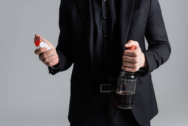 обрезанный вид человека с пачкой сигарет и бутылкой виски, изолированной на сером - Фото, изображение