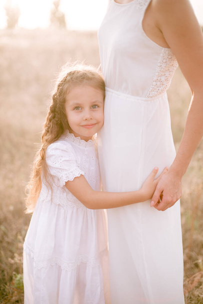 A cute little girl with long blond curly hair and her mother in a white summer dress and a straw boater hat in a field in the countryside in summer at sunset. Nature and Ecolife - Foto, afbeelding