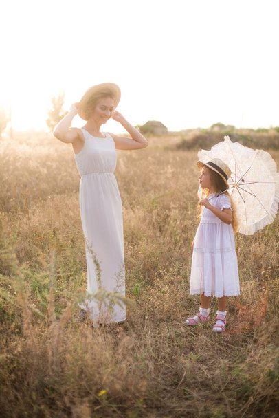 A cute little girl with long blond curly hair and her mother in a white summer dress and a straw boater hat in a field in the countryside in summer at sunset. Nature and Ecolife - Foto, Imagem