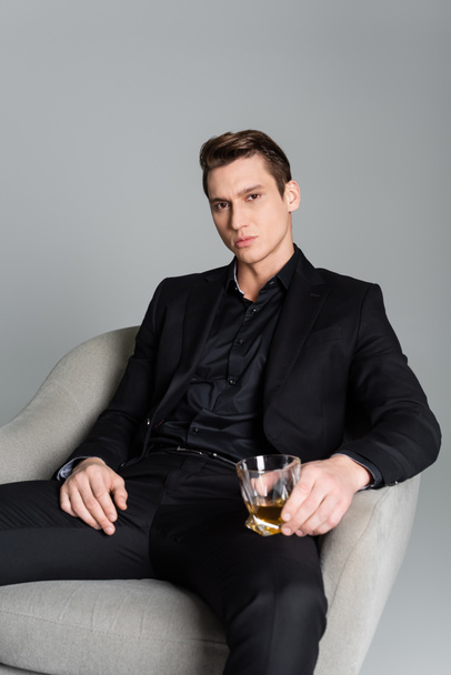 serious man in black suit sitting in armchair with glass of whiskey isolated on grey - Foto, imagen