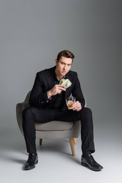 longitud completa del hombre en traje negro sentado en sillón con dinero y whisky sobre fondo gris - Foto, Imagen