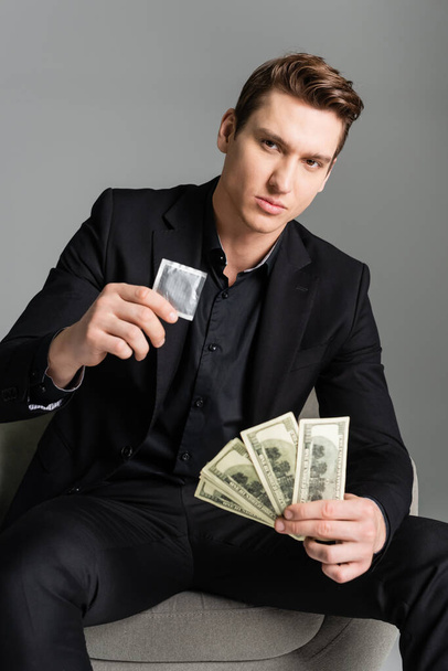 confident man holding condom and dollars while looking at camera isolated on grey - Zdjęcie, obraz