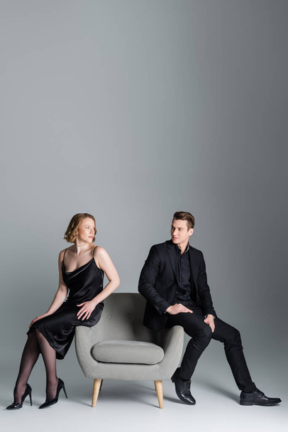 full length of couple in black clothes looking at each other on armchair on grey background - Φωτογραφία, εικόνα