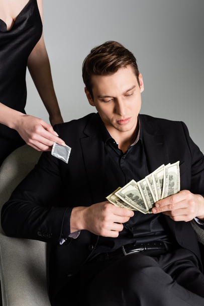 мужчина в черном костюме считает деньги рядом с женщиной с презервативом, изолированным на сером - Фото, изображение