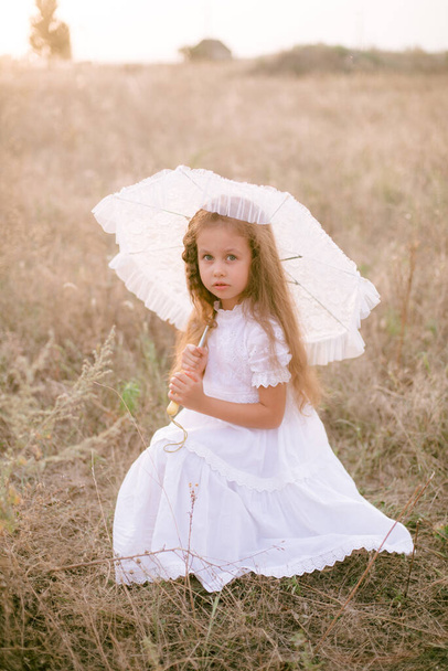 A cute little girl with long blond curly hair in a white summer dress and a straw boater hat with a white lace umbrella in a field in the countryside in summer at sunset. Nature and Ecolife - Photo, Image