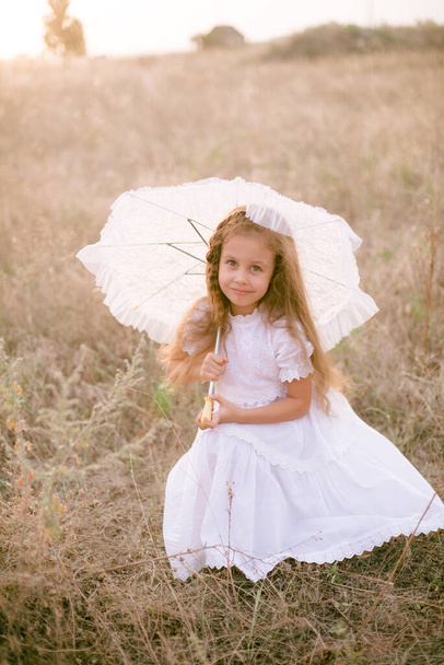 A cute little girl with long blond curly hair in a white summer dress and a straw boater hat with a white lace umbrella in a field in the countryside in summer at sunset. Nature and Ecolife - Photo, image