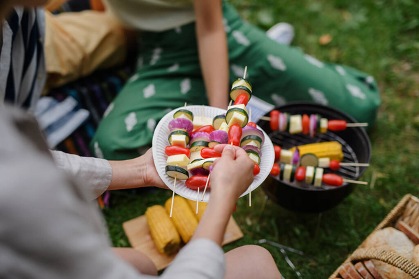 Close-up of young friends putting corn and skewers on grill and having barbecue when camping in campground. - 写真・画像