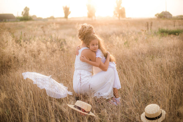 A cute little girl with long blond curly hair and her mother in a white summer dress and a straw boater hat in a field in the countryside in summer at sunset. Nature and Ecolife - Zdjęcie, obraz