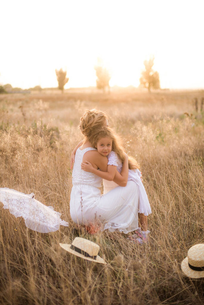 A cute little girl with long blond curly hair and her mother in a white summer dress and a straw boater hat in a field in the countryside in summer at sunset. Nature and Ecolife - Φωτογραφία, εικόνα