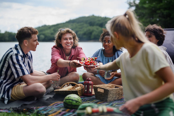 A group of multiracial young friends camping near lake and and having barbecue together, looking at camera. - Фото, изображение