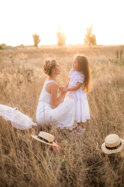 A cute little girl with long blond curly hair and her mother in a white summer dress and a straw boater hat in a field in the countryside in summer at sunset. Nature and Ecolife - Фото, изображение