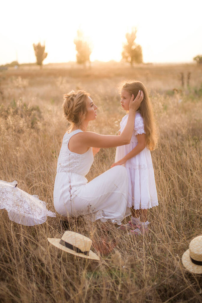 A cute little girl with long blond curly hair and her mother in a white summer dress and a straw boater hat in a field in the countryside in summer at sunset. Nature and Ecolife - Photo, image