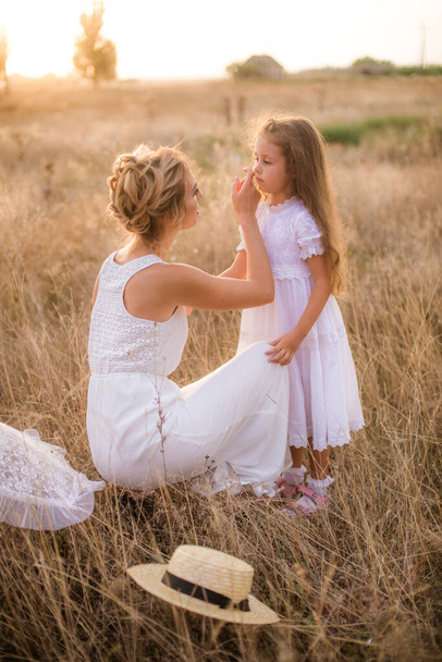 A cute little girl with long blond curly hair and her mother in a white summer dress and a straw boater hat in a field in the countryside in summer at sunset. Nature and Ecolife - Foto, immagini