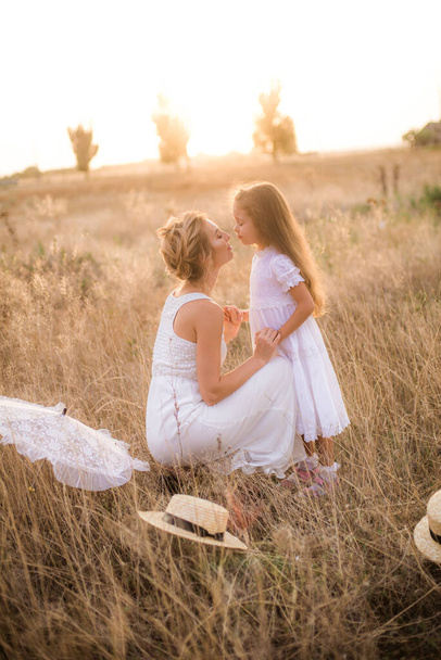 A cute little girl with long blond curly hair and her mother in a white summer dress and a straw boater hat in a field in the countryside in summer at sunset. Nature and Ecolife - Fotó, kép