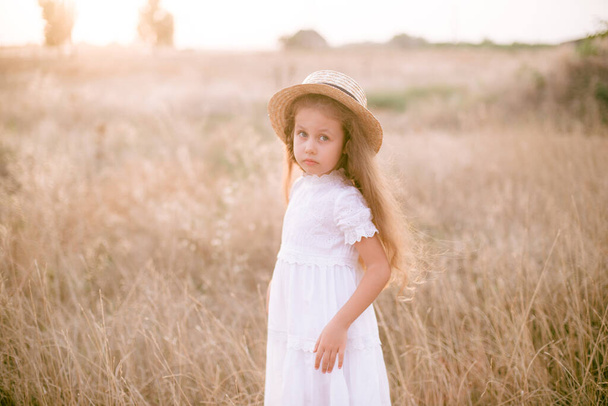 A cute little girl with long blond curly hair and her mother in a white summer dress and a straw boater hat in a field in the countryside in summer at sunset. Nature and Ecolife - Фото, зображення