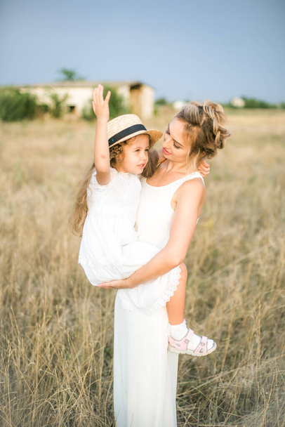 A cute little girl with long blond curly hair and her mother in a white summer dress and a straw boater hat in a field in the countryside in summer at sunset. Nature and Ecolife - Photo, Image