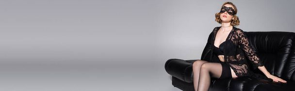 mladá a sexy žena v krajkové masce a černé spodní prádlo sedí na koženém gauči izolované na šedé, banner - Fotografie, Obrázek