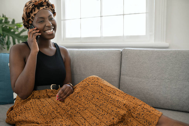 Прекрасна молода чорношкіра жінка з Африки, яка сидить на дивані вдома, розмовляє розумним телефоном, посміхається, стріляє з копіювального простору. - Фото, зображення