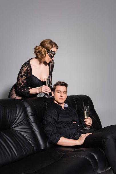 соблазнительная женщина с бокалом шампанского рядом с мужчиной на черном кожаном диване, изолированном на сером - Фото, изображение