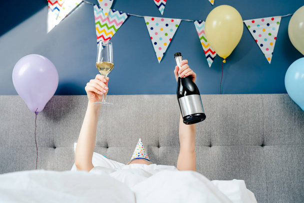 Hands of a woman with a bottle and glass of champagne lying on the bed in the bedroom decorated with balloons and garland.Time for yourself Celebration at home. Happy birthday concept. Selective focus - Foto, Bild