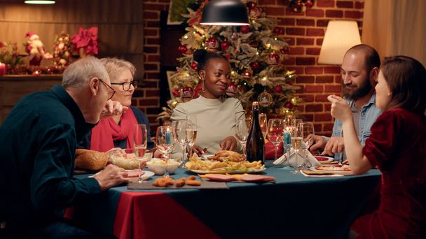 Membros da família festiva feliz apreciando o jantar de Natal em casa enquanto comem juntos. Pessoas multiétnicas alegres na sala de jantar celebrando férias de inverno com comida caseira. Tiro portátil - Foto, Imagem