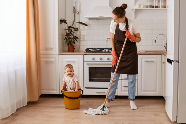 Indoor shot of attractive woman making domestic work and taking care of her infant baby, wiping washing floor in kitchen, looking at her child with smile. - Photo, Image