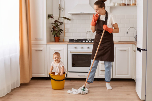 Full length shot of nice attractive hardworking woman making fast domestic work with her cute baby daughter in bucket, wiping floor in modern light white interior kitchen. - Φωτογραφία, εικόνα