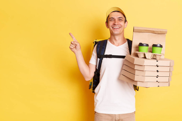 Délicieux livreur souriant heureux portant t-shirt et casquette tenant boîtes à pizza et boisson à emporter, posant isolé sur fond jaune, pointant doigt de côté à l'espace de copie pour la promotion. - Photo, image