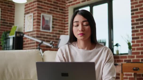 Focused freelancer woman working remotely from home on modern laptop. Beautiful young adult person doing remote freelancing work on portable computer while sitting on sofa inside apartment. - Footage, Video