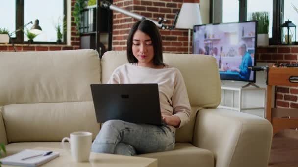 Soustředěná mladá dospělá žena pracující na dálku na notebooku, zatímco sedí na pohovce v obývacím pokoji doma. Atraktivní asijský freelancer dělá vzdálenou práci na moderním přenosném počítači. - Záběry, video