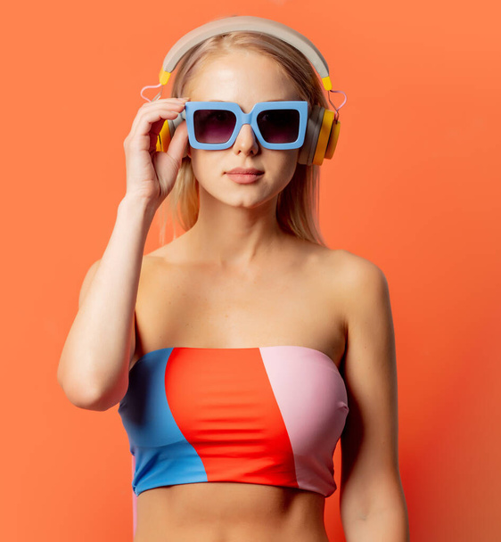 Mulher loira elegante em terno de natação colorido com fones de ouvido e óculos de sol no fundo laranja - Foto, Imagem