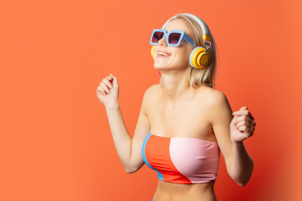 Stylish blond woman in colored swimming suit with headphones and sunglasses on orange background - Foto, Imagem