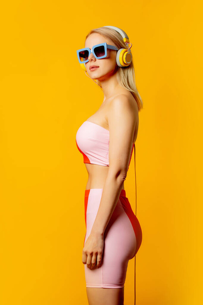 Stijlvolle blonde vrouw in gekleurd zwempak met hoofdtelefoon en zonnebril op gele achtergrond - Foto, afbeelding