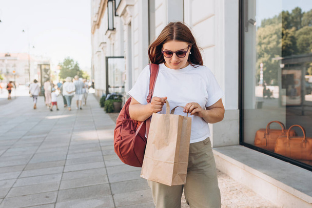 Piękna, modna młoda kobieta w okularach przeciwsłonecznych z torbą na zakupy stojącą na ulicy miasta, czarny piątek. Koncepcja miejskiego stylu życia. Transport przesyłek. - Zdjęcie, obraz