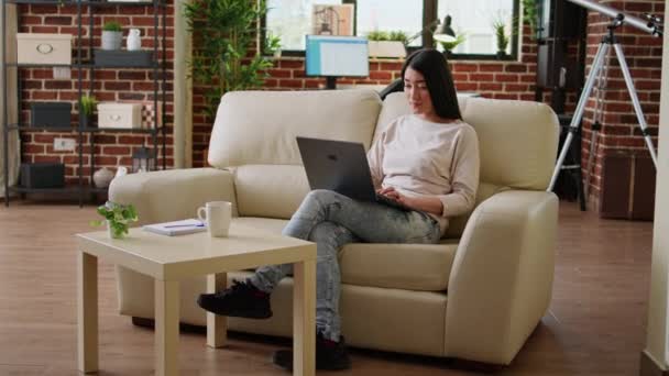 Επικεντρωμένος νεαρός ενήλικος ασιάτης φοιτητής που παρακολουθεί μαθήματα online ενώ πληκτρολογεί σημαντικά μαθήματα στον σύγχρονο φορητό υπολογιστή στο σπίτι. Όμορφη γυναίκα κάθεται στον καναπέ, ενώ εργάζονται εξ αποστάσεως σε φορητό υπολογιστή. - Πλάνα, βίντεο