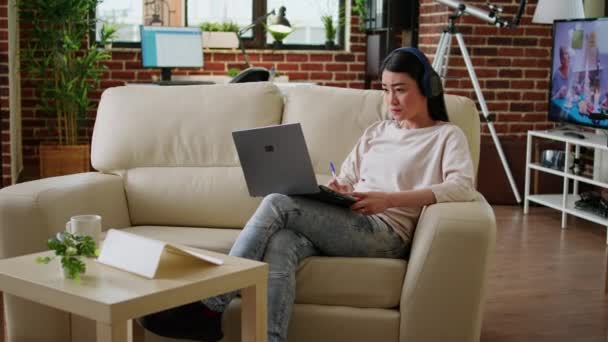 Focused young adult student taking notes while attending online class on laptop at home. Concentrated asian freelancer working remotely on portable computer while sitting on sofa inside apartment. - 映像、動画