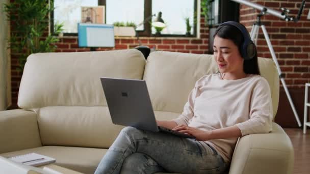 Uśmiechnięta uczennica uczęszczająca na zajęcia online na laptopie siedząc na sofie w domu. Piękna młoda kobieta pracuje zdalnie na przenośnym komputerze wewnątrz mieszkania podczas pracy na własny rachunek. - Materiał filmowy, wideo