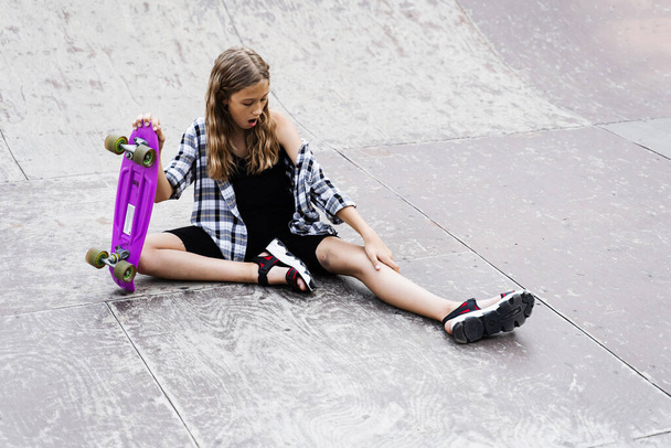 Actief kind meisje na de val van penny board gewond, zitten en kijken naar blauwe plekken op de sportbaan op skate park speeltuin - Foto, afbeelding