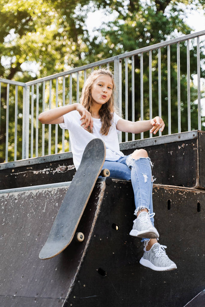 Having fun with skate board. Funny child girl with skate sitting on sport ramp, smiling and grimacing on skate playground. Active teenager posing with skate board. Extreme lifestyle - Photo, image