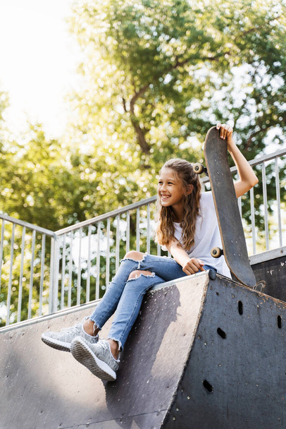 Happy smiling girl with skate board sitting on skate playground and having fun. Extreme sport lifestyle. Laughing child with skate board posing on sport ramp - Foto, Imagem