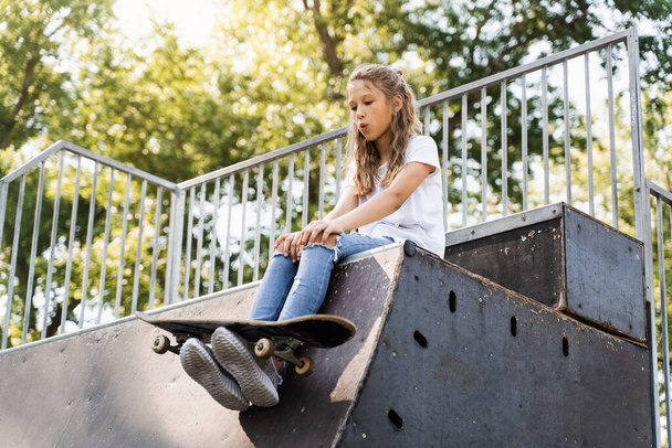 Happy smiling girl with skate board sitting on skate playground and having fun. Extreme sport lifestyle. Laughing child with skate board posing on sport ramp - Fotografie, Obrázek