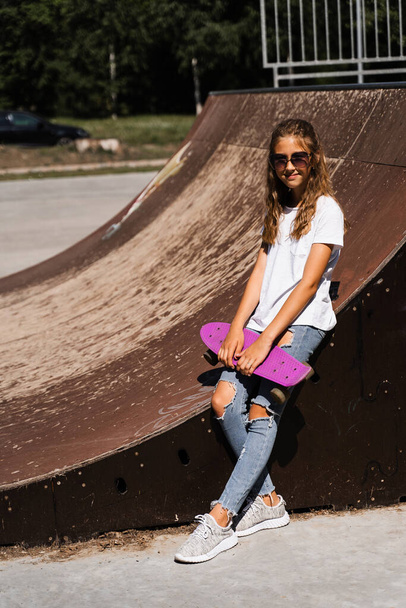 Ragazza di moda bambino in occhiali in posa con penny board sul parco giochi skate. Adolescente attivo con attrezzatura sportiva sulla rampa sportiva. Stile di vita estremo - Foto, immagini