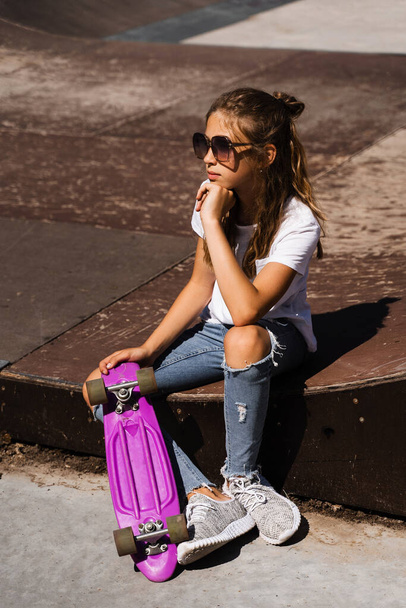 Kind mit Groschenbrett. Ein junges Mädchen mit Brille und Skateboard sitzt auf einer Sportrampe auf dem Skaterplatz. Extremer Lebensstil. Kreative Werbung für Skate- oder Penny-Boardshops oder Geschäfte - Foto, Bild