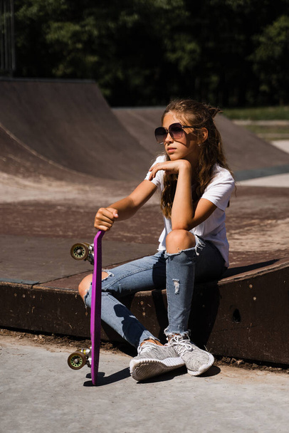 Child with penny board. Young girl teenager in glasses with skate board sitting on sport ramp on skate playground. Extreme lifestyle. Creative advert for skate or penny board shops or stores - Photo, Image