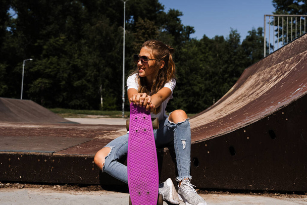 Счастливая улыбчивая девочка с доской для скейта, сидящая на детской площадке и развлекающаяся. Экстремальный спорт. Смеющийся ребенок со скейтбордом позирует на спортивной рампе - Фото, изображение